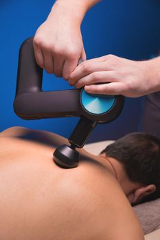 Medium shot of caucasian professional male massage therapist getting back muscles with massage gun impact tool of muscular athlete, in spa treatments, lying on massage table