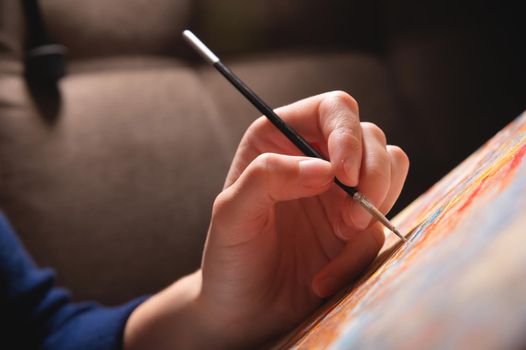 Close-up of a young female artist's hand with a thin brush painting a picture on canvas in a dark room. Shallow depth of field. high contrast.