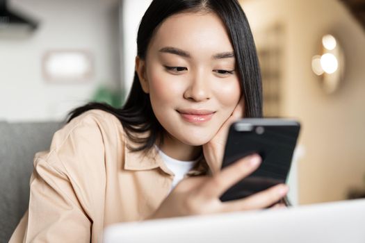Portrait of asian girl sitting with laptop, checking her phone and smiling, browsing websites on smartphone.