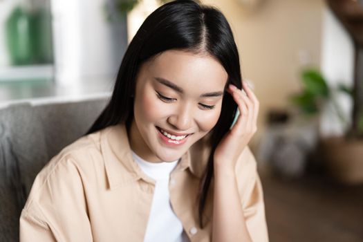 Cute asian girl student smiling, looking down and tucking hair behind ear, sitting at home indoors in casual clothes.