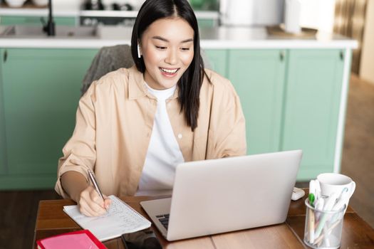 Young asian woman taking notes, working during video conference with laptop at home.