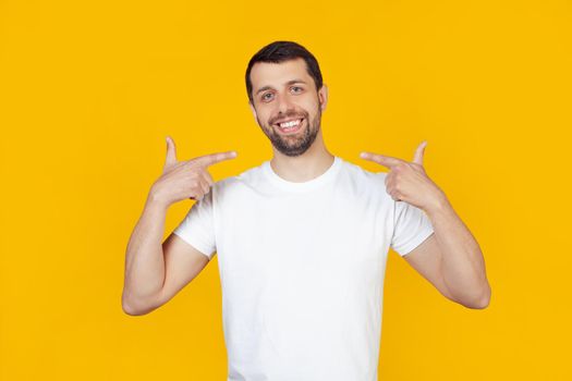 Young man with a beard in a white t-shirt happy face smiling confidently showing and pointing with fingers teeth and mouth. Health concept. stands on isolated yellow background.
