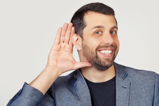 Young businessman man with a beard in a jacket, smiling with his hand above his ear, listening to rumors or gossip. Deafness concept. Portrait of a man on a gray background.