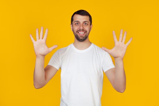 Modern young man with a beard in a white tank top shows number nine with fingers on hand smiling confidently and happily, looking into the camera The man shows ten fingers. Number 10 yellow background