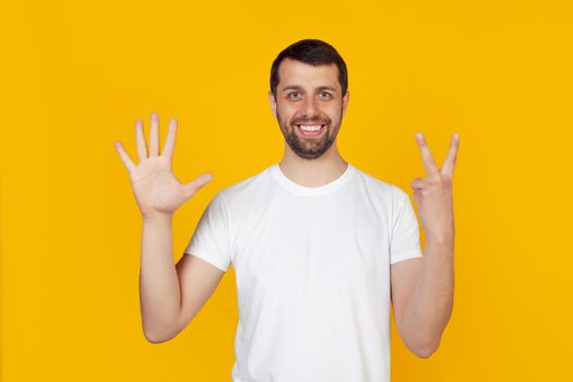Modern young man with a beard in a white tank top shows number seven with fingers on hand, smiling confidently and happily, looking into the camera. The man shows seven fingers. Number 7.