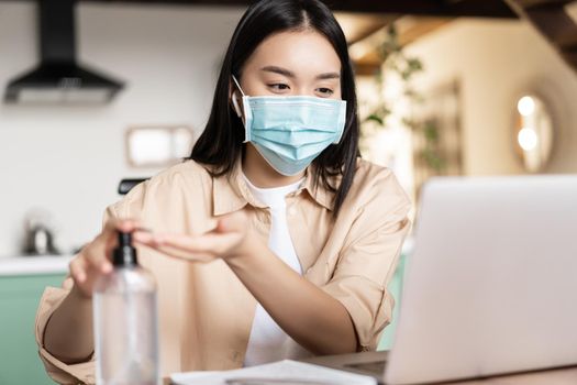 Young asian woman in medical face mask sitting at coworking space, attend online lecture or webinar on laptop and using hand sanitizer to wash hands.