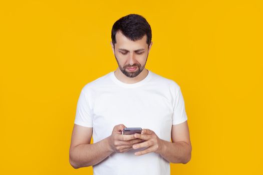 A young man with a beard in a white T-shirt holds in his hand and looking into a smartphone looking for information on the Internet, a natural expression, stands on an isolated yellow background.