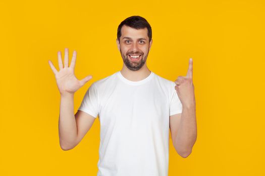 Modern young man with a beard in a white tank top shows number six with fingers on hand, smiling confidently and happily, looking into the camera The man shows six fingers. Number 6. yellow background