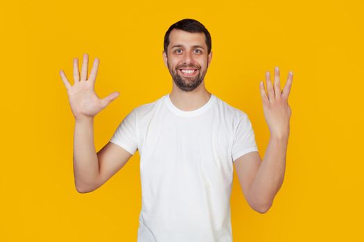Modern young man with a beard in a white tank top shows number nine with fingers on hand, smiling confidently and happily looking into the camera The man shows nine fingers. Number 9 yellow background