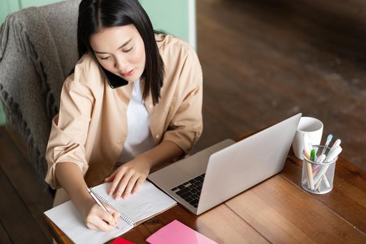 Upper angle shot of asian woman studying on phone, talking on mobile phone and writing down notes. Working girl using laptop and cellphone, talks to customer.