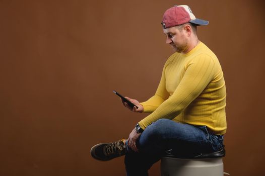Chubby caucasian businessman sitting uses smart mobile phone while looking at camera. Studio portrait of a casual man sitting with a smartphone in his hands.