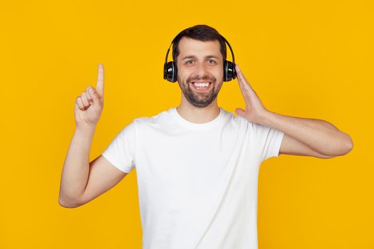 Young man with a beard in a white t-shirt listens to music with headphones over isolated yellow background, surprised with an idea or question, pointing with a finger with a happy face, number one.