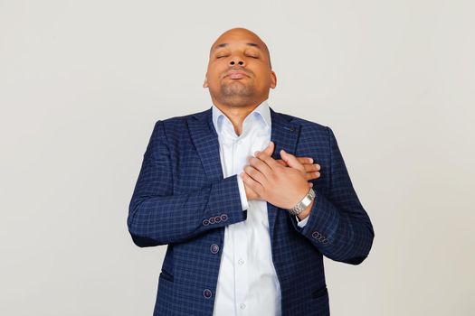 Portrait of grateful young African American businessman guy smiling with hands on chest with closed eyes and grateful gesture on face. Health concept. Standing on a gray background.