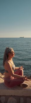 Female in sports clothes sitting by the beautiful seashore in lotus position and training on sunny day