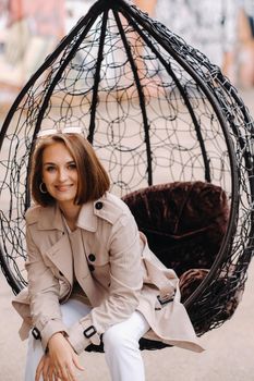 A happy stylish girl In a gray coat is sitting outside in an armchair.