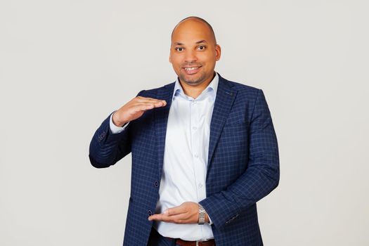 Portrait of happy young african american businessman guy gesturing with hands showing big and large size sign, symbol of measure. Smiling looking into the camera. Standing on a gray background