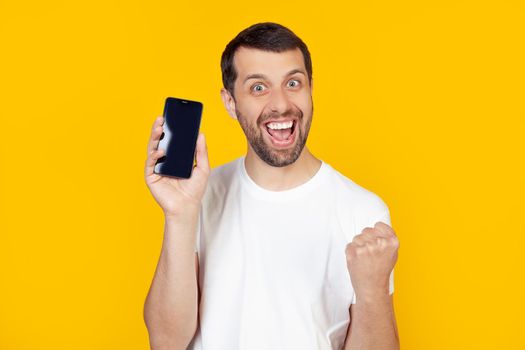A young man with a beard in a white T-shirt shows a smartphone screen, shouts with pride and celebrates victory and success, is very excited, rejoices in emotions. Stands on isolated yellow background.