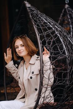 A happy stylish girl In a gray coat is sitting outside in an armchair.