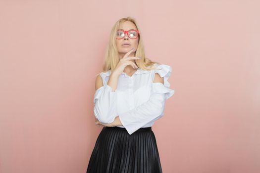 Beauty, fashion portrait. Elegant business style. Portrait of a beautiful blonde woman in white blouse and black skirt posing at studio on a pink background. Wearing red glasses