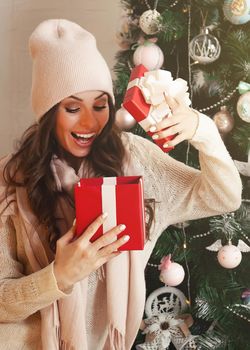 Smiling and happy, beautiful young woman in winter clothes with a red Christmas gift box on the background of a decorated Christmas tree. The girl delightedly opens the gift, greets the New Year.
