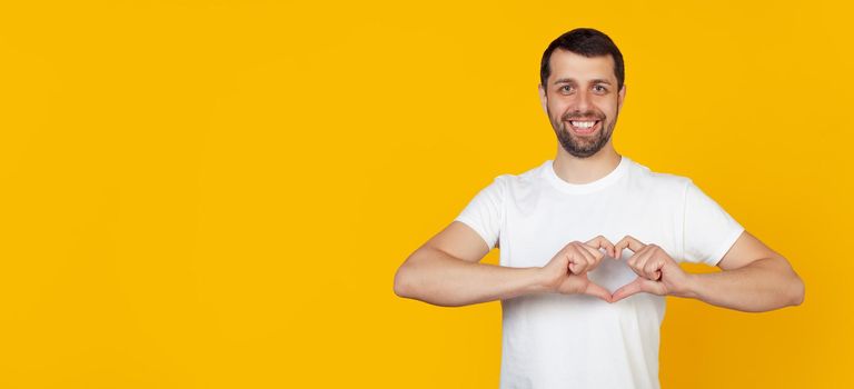 Handsome young man, a guy with a beard in a white T-shirt, smiles and makes a heart-shaped symbol with his finger, expresses love and a positive romantic feeling. Yellow background