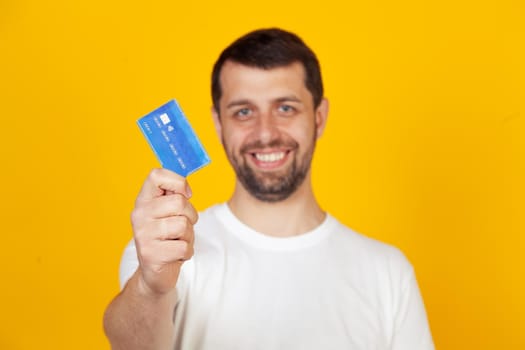Young man with a beard in a white T-shirt holding a credit card with a happy face, stands and smiles with a confident smile, showing teeth. Stands on isolated yellow background.
