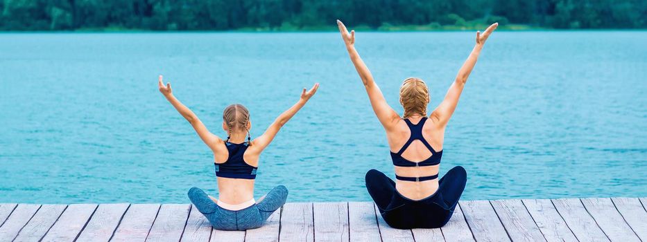 Mother and daughter doing yoga at the shore of the lake in summer.