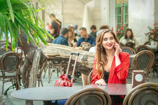 Young cute girl is talking by mobile phone sitting alone in coffee shop outdoors.