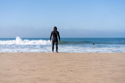 One man dressed black wetsuit standing ocean beach rear view looking on horizon. Male person surfer enjoy sea view