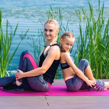 Portrait of mother and daughter sitting on yoga mat by the lake outdoors. Healthy and exercise concept.