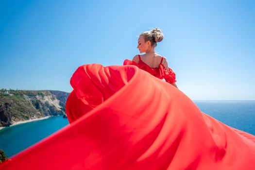 Blonde with long hair on a sunny seashore in a red flowing dress, back view, silk fabric waving in the wind. Against the backdrop of the blue sky and mountains on the seashore