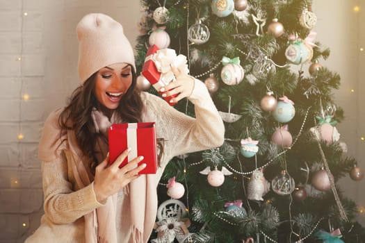 Smiling and happy, beautiful young woman in winter clothes with a red Christmas gift box on the background of a decorated Christmas tree. The girl delightedly opens the gift, greets the New Year.