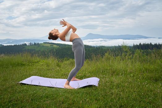 Active young woman in sport outfit practicing yoga during free time on fresh air. Female person enjoying beautiful view on mountains during outdoors workout.