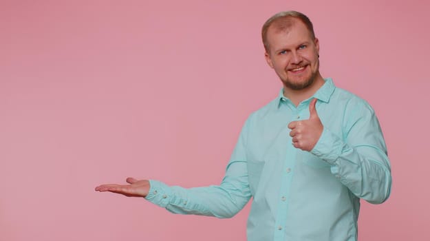 Handsome one bearded man showing thumbs up and pointing empty place, advertising area for commercial text, copy space for goods promotion. Young adult guy. Studio shot indoors on pink wall background