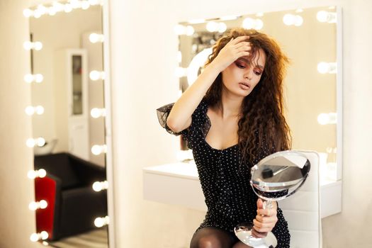 Beautiful girl model brunette, looks in the mirror straightens her hair in a beauty salon. Young woman in black dress looking in the mirror at herself getting ready for a date.