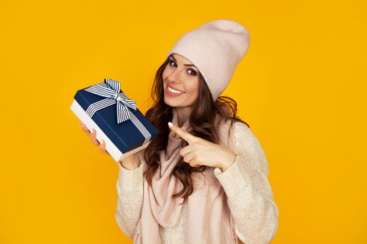 Happy young woman smiling pointing finger holding blue gift box in hand. A gift to your beloved man. A girl in a sweater looks at the camera and points to a gift. Christmas New Year holiday concept