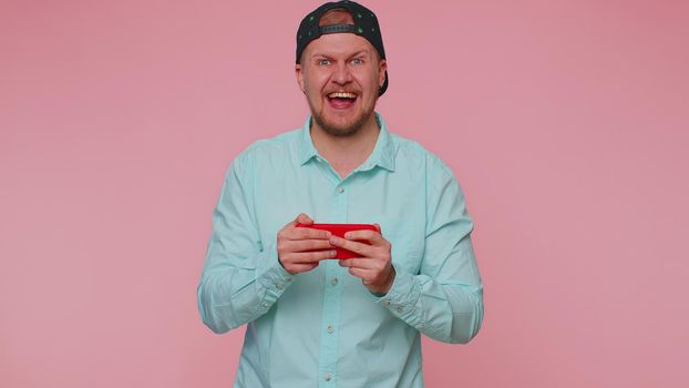 Worried funny addicted man in shirt enthusiastically playing racing video games on mobile phone. Young adult guy using smartphone gadget app with drive simulator isolated on pink studio background
