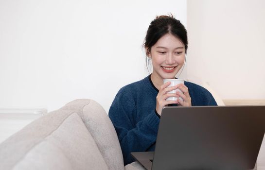 Portrait beautiful young asian woman Working on Laptop Online, Using Internet and hold the cup. Sitting on Sofa at Home, Free Space.