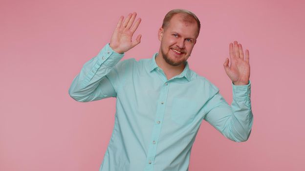 Handsome friendly man in shirt smiling friendly at camera and waving hands gesturing hello or goodbye, welcoming with hospitable expression. Young happy guy indoors isolated on pink wall background