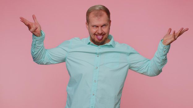 Funny joyful sincere adult man 30 years old in shirt making playful silly facial expressions and grimacing, fooling around showing tongue. Young handsome guy isolated alone on pink wall background