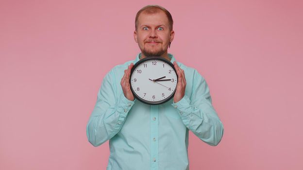Joyful young man holding clock watch, hiding, checking time on clock, running late to work, being in delay, deadline on pink studio background. Serious guy looking at camera worrying to be punctual