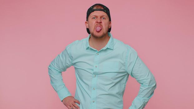 Cheerful funny man in blue shirt showing tongue making faces at camera, fooling around, joking, aping with silly face, teasing. Young adult blonde guy boy isolated alone on pink studio wall background
