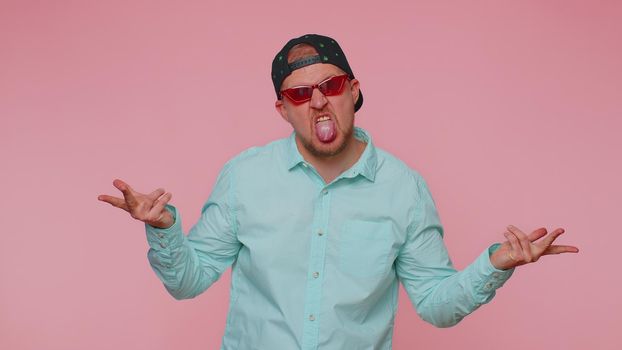 Funny joyful sincere man in sunglasses and marihuana cap making playful silly facial expressions and grimacing, fooling around showing tongue. Young handsome guy isolated alone on pink wall background