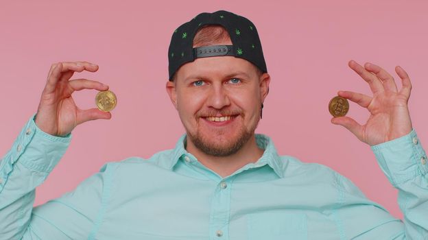 Happy man winner showing golden bitcoins. Career wealth, cryptocurrency investment, mining future technology. Young adult guy fooling making faces. Happy businessman isolated on pink studio background