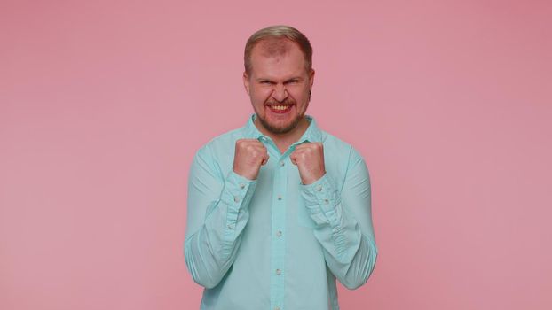 Happy joyful man in casual blue shirt shouting, raising fists in gesture I did it, celebrating success, winning, birthday, lottery goal achievemen. Adult guy boy indoors on pink studio wall background