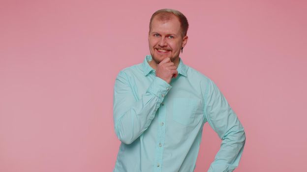 Cheerful lovely bearded adult man in casual blue shirt smiling and looking at camera. Young fashion model guy boy with gray eyes indoor studio shot isolated on pink background. Male nature beauty