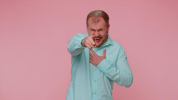 Amused man in casual blue shirt pointing finger to camera, laughing out loud, taunting making fun of ridiculous appearance, funny joke. Young guy boy posing alone on pink studio wall background