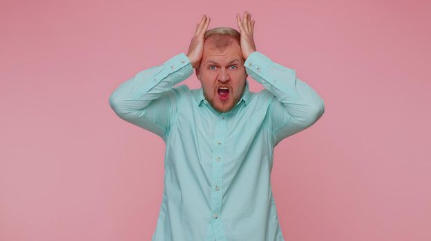 Man in blue shirt screams from stress tension problems feels horror hopelessness fear surprise shock expresses gestures rage. Young adult blonde guy boy isolated alone on pink studio wall background