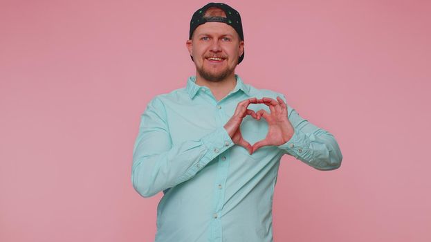 Man in love. Smiling man 30s in blue shirt makes heart gesture demonstrates love sign expresses good feelings and sympathy. Young adult blonde guy boy isolated alone on pink studio wall background