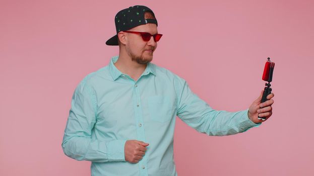 Man in blue shirt traveler blogger taking selfie on mobile phone selfie stick, communicating video call online with subscribers. Young adult guy boy 30s isolated alone on pink studio wall background